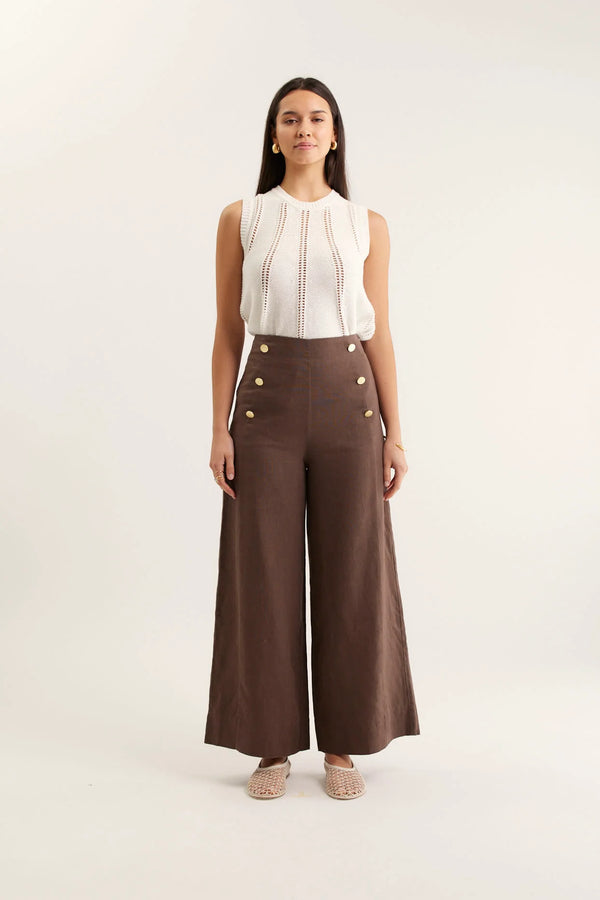 Woman wearing a white sleeveless top and brown wide-leg pants with gold buttons on a white background