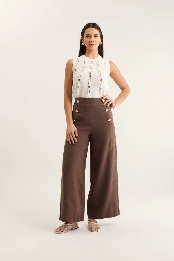 Woman wearing a white sleeveless top and brown wide-leg pants with buttons on a white background