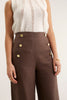 Person wearing a white sleeveless top and brown pants with gold buttons on a plain background