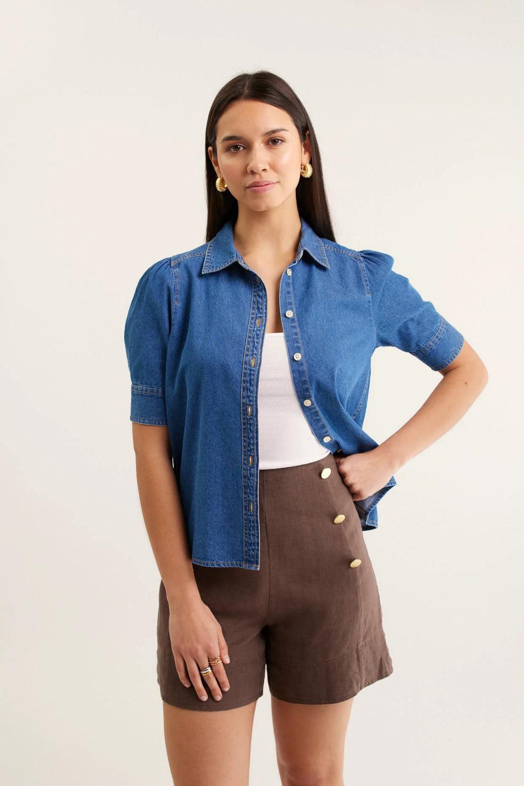 Woman wearing a blue denim shirt over a white top and brown shorts on a light background