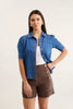 Woman wearing a blue denim shirt over a white top and brown shorts on a light background