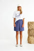 Woman wearing a white t-shirt with a horse logo and blue shorts standing next to a stack of hay.
