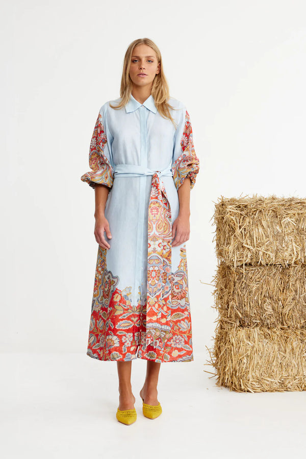 Woman wearing a light blue dress with a colorful patterned kimono-style jacket, standing next to a stack of hay.