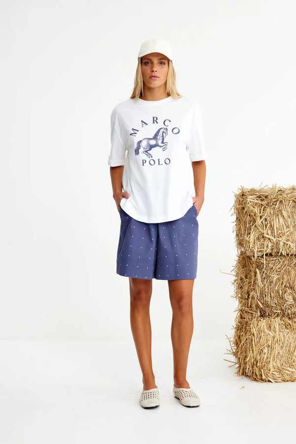 Person wearing a white t-shirt with branding and blue shorts standing next to a stack of hay.