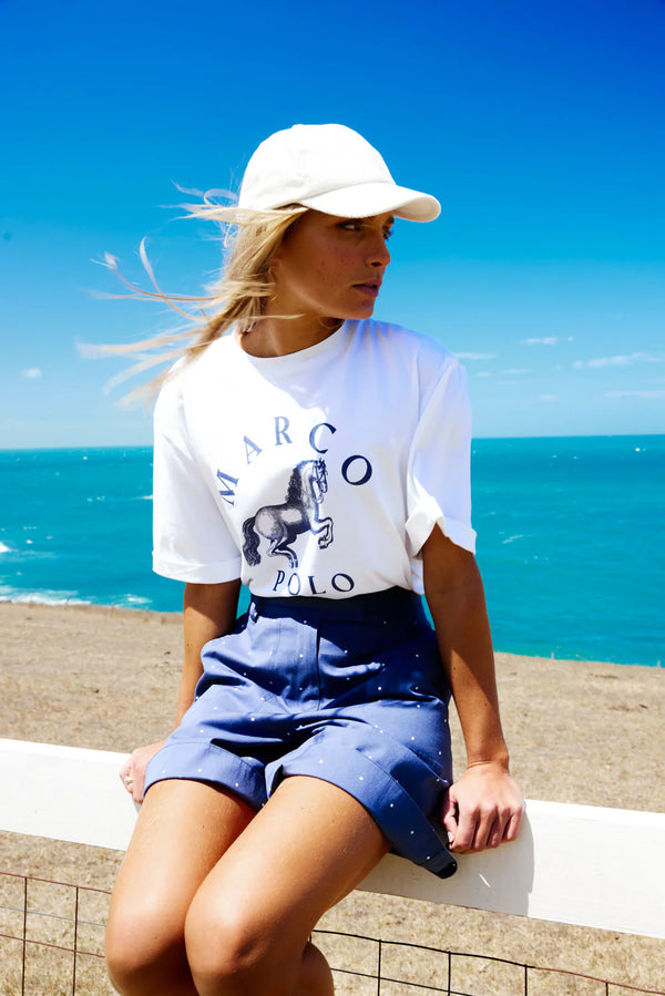 Person wearing a white t-shirt with 'Marc O' Polo' logo and blue shorts sitting by the ocean.