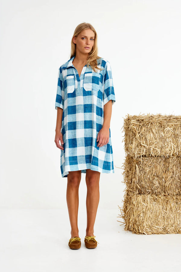 Woman wearing a blue and white checkered dress standing next to a bale of hay on a white background