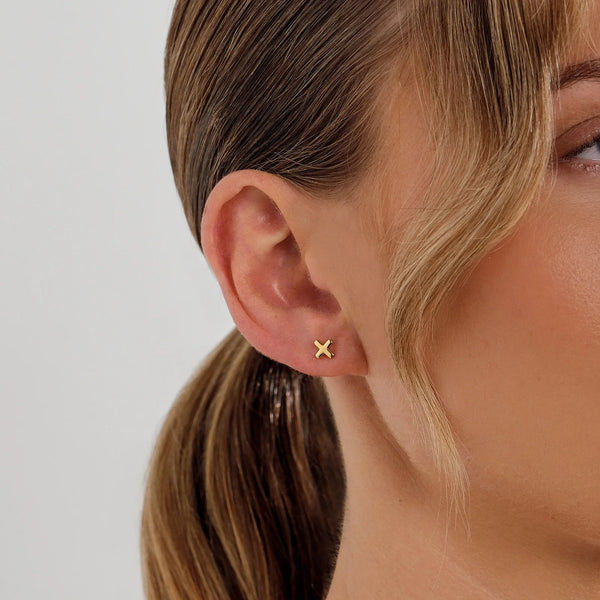 Close-up of an ear wearing a gold earring with a 'X' design on a neutral background